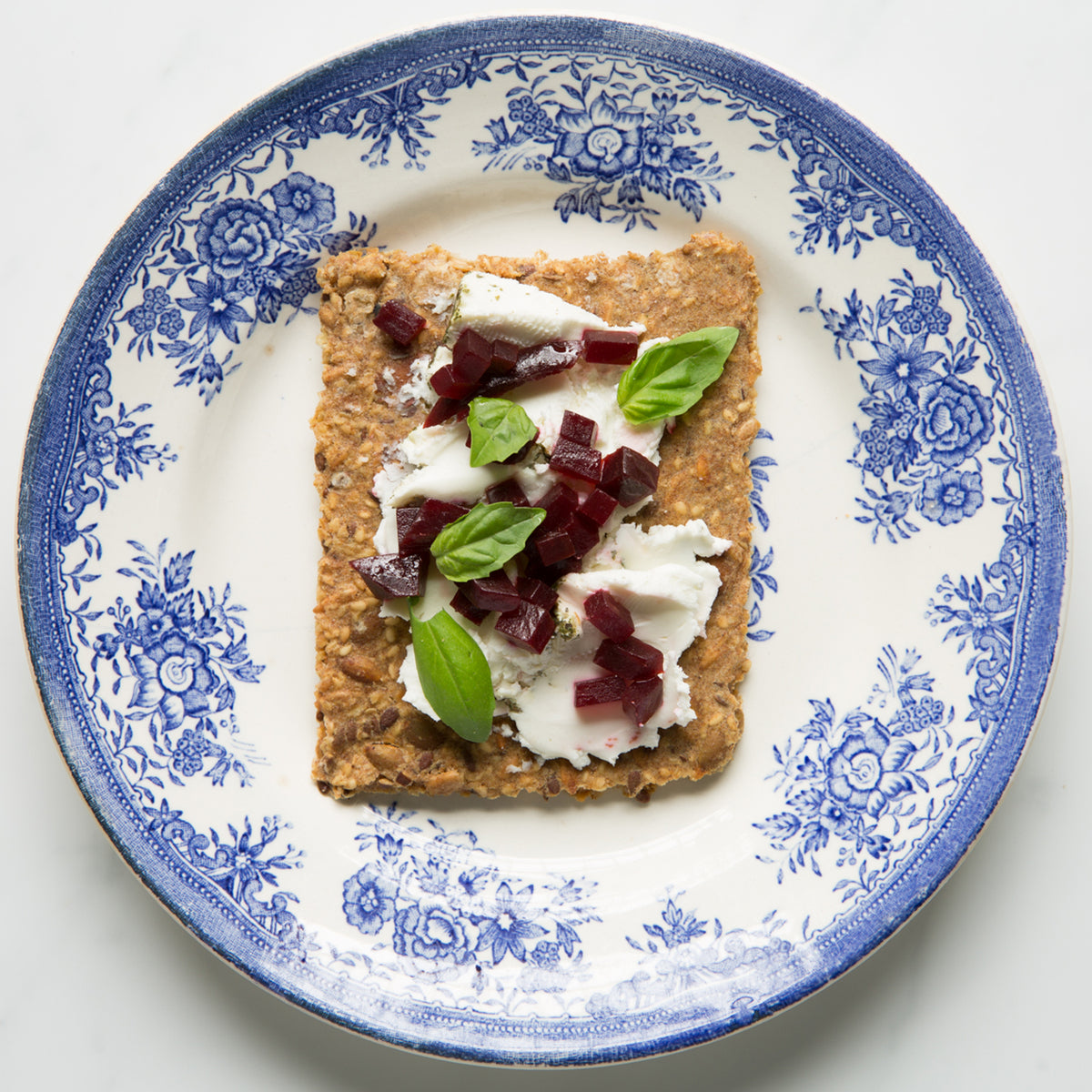 Overhead view of Rosemary Lemon Knekkebrød with cream cheese, beets, and basil on a decorative blue and white plate.