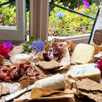 Assorted meats, soft cheeses, and Rosemary and Lemon Knekkebrød  on a wooden board with flowers and a window in the background.