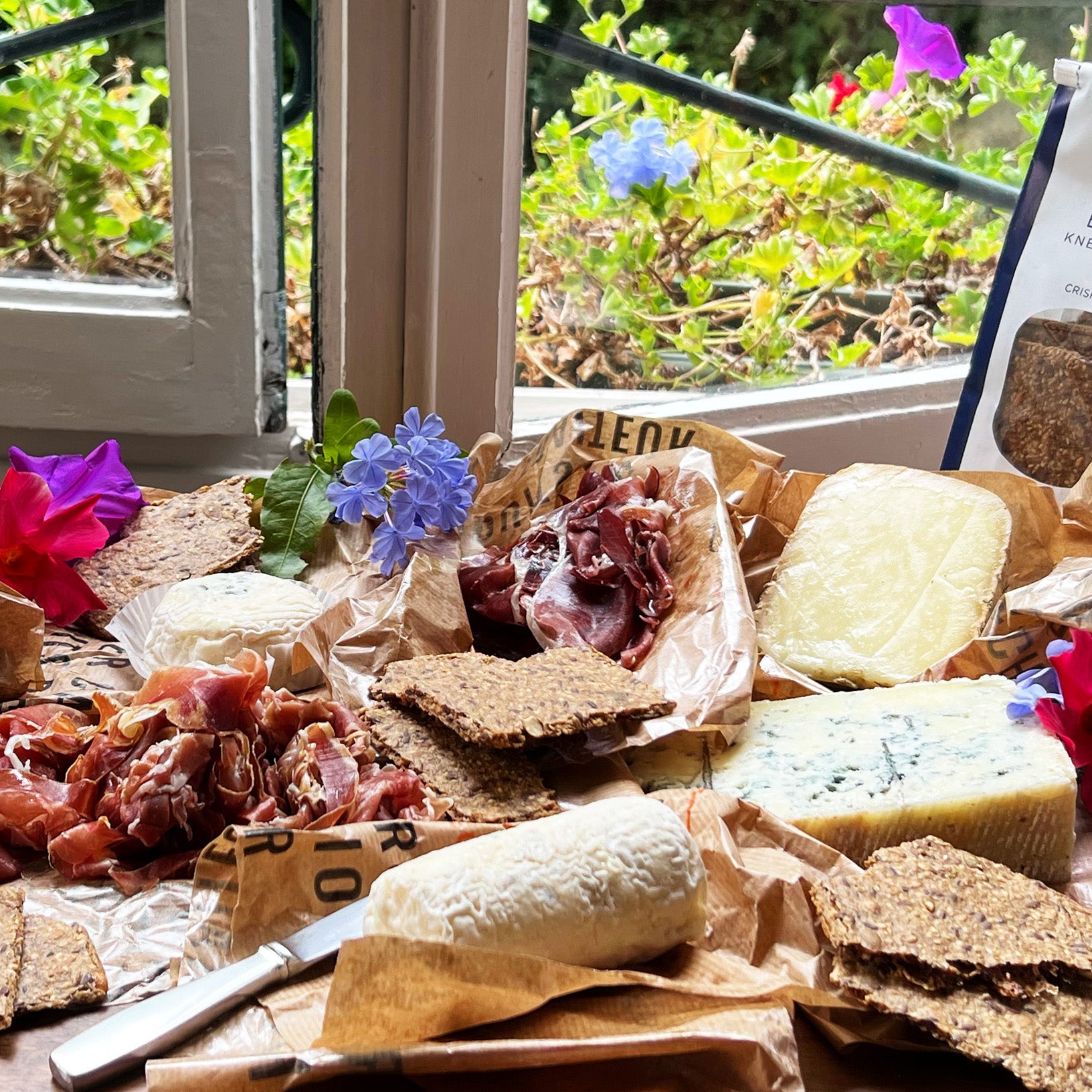 Assorted meats, soft cheeses, and Rosemary and Lemon Knekkebrød  on a wooden board with flowers and a window in the background.
