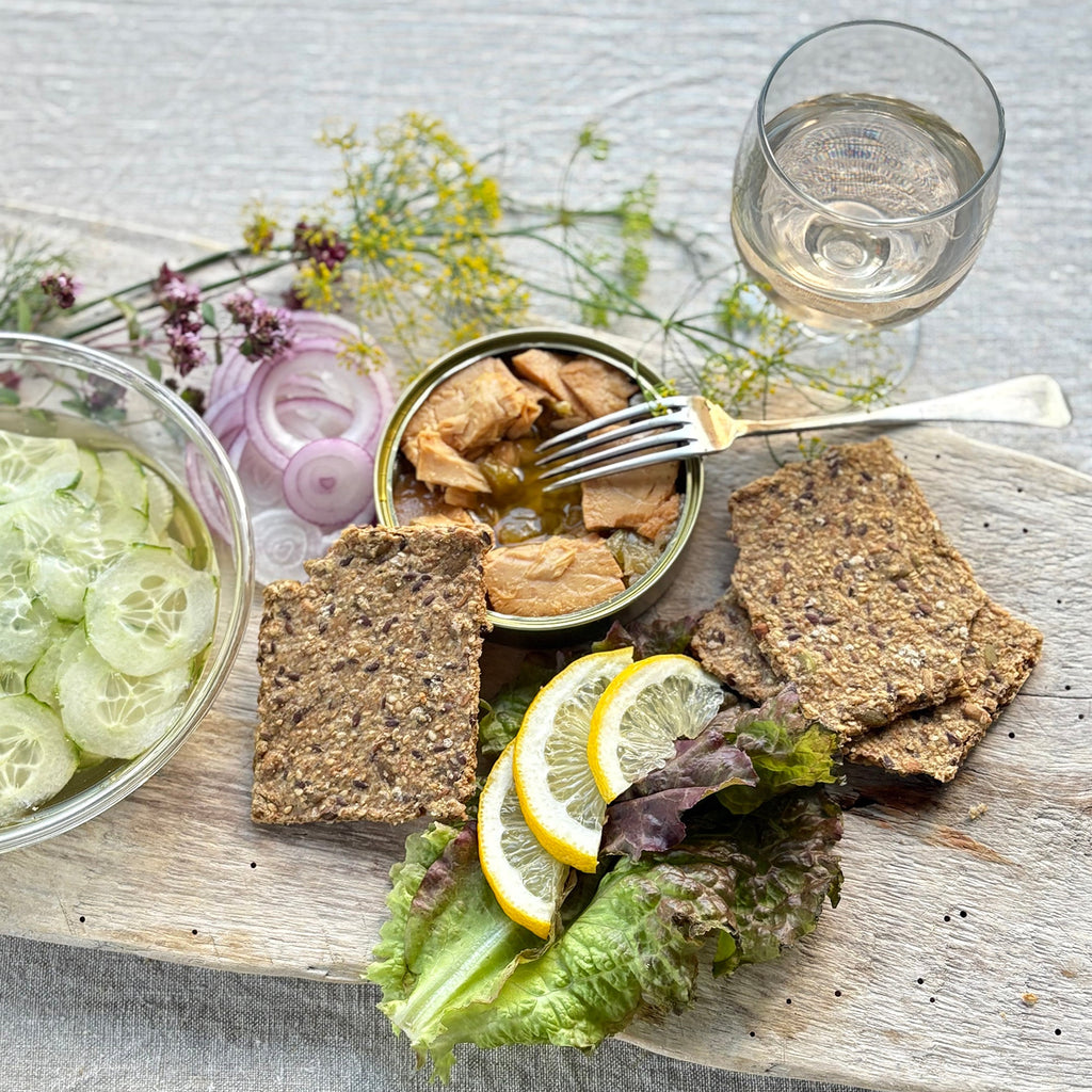 Platter with Sea Salt Flakes Crispbread, pickled cucumbers, lemon slices, and a can of fish on a light surface.