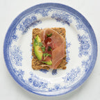 Small square of Sea Salt Flakes crispbread with avocado and prosciutto on a decorative plate.