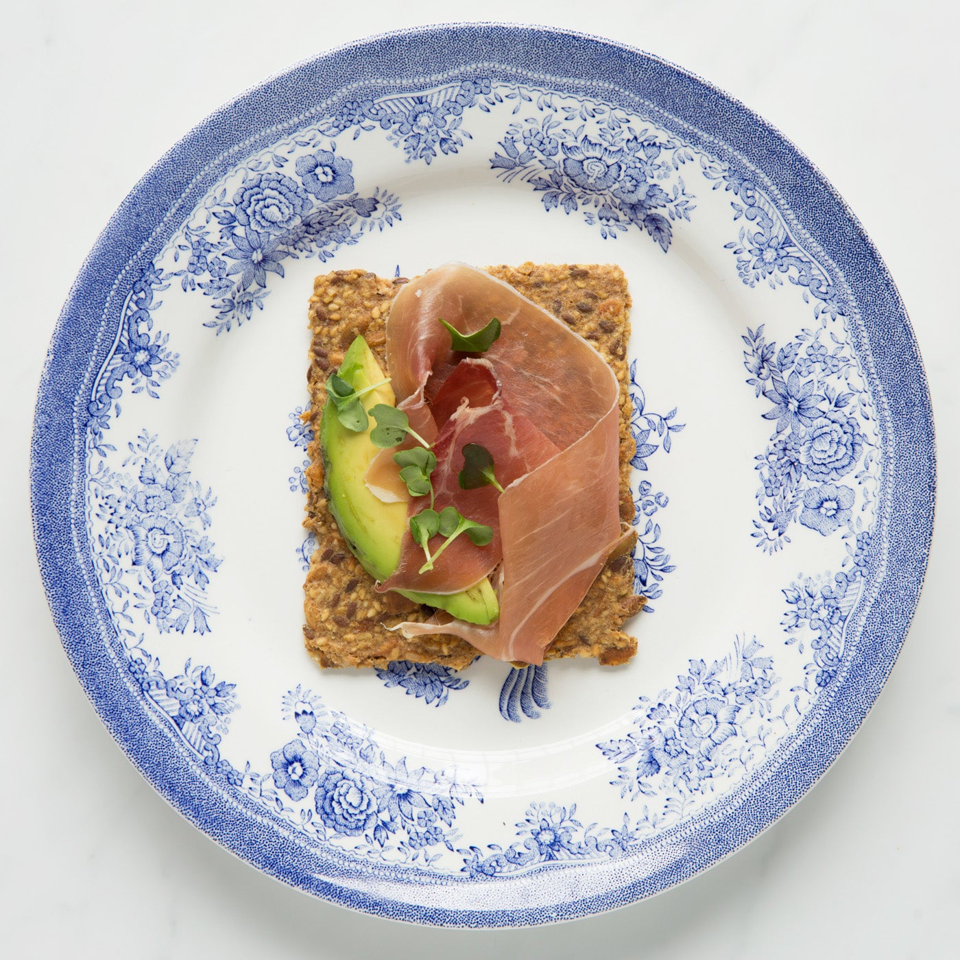 Small square of Sea Salt Flakes crispbread with avocado and prosciutto on a decorative plate.