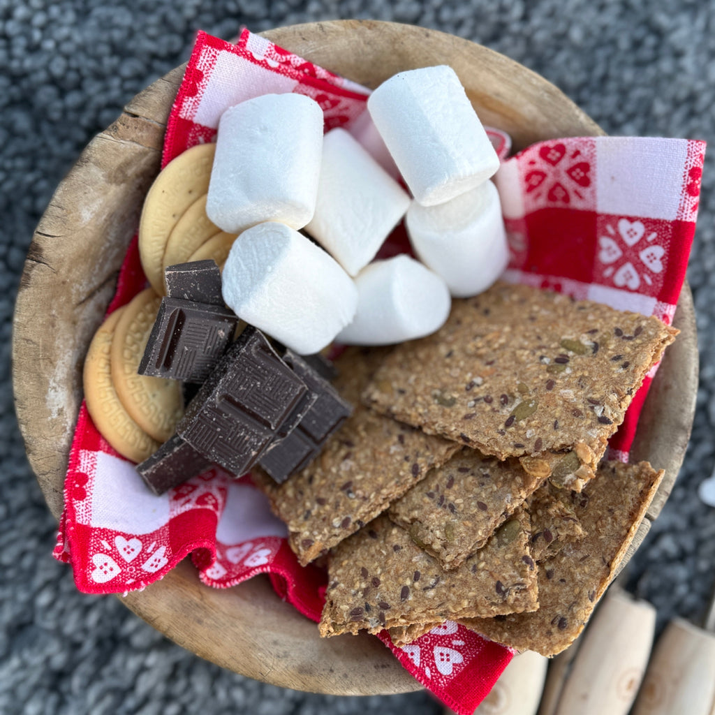 Wooden bowl with Sea Salt Flakes Knekkebrød, chocolate bars, and marshmallows on a red checkered cloth.